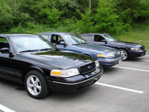 1997 Mercury Grand Marquis - Three dark-colored sedans are parked in a row on an asphalt surface with white parking lines. The closest vehicle is black, followed by two…