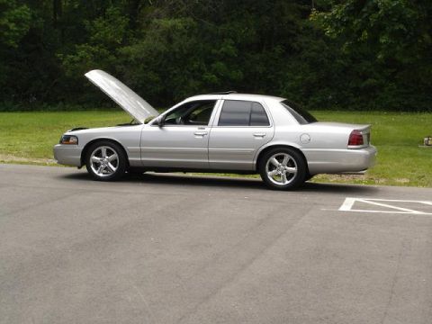 1997 Mercury Grand Marquis - A silver four-door sedan with the hood open is parked on the side of a paved road with a grassy area and trees in the background.