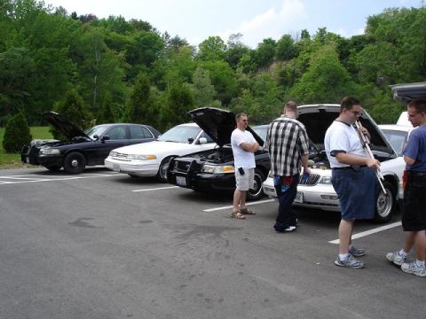 1997 Mercury Grand Marquis - A line of four sedans, three with hoods open, are parked in a lot with a wooded hillside in the background. Several men stand around the ve…