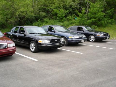 1997 Mercury Grand Marquis - Three sedans are parked in a lot with a wooded area in the background. The closest vehicle is a dark red sedan, followed by a black sedan a…