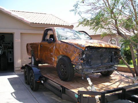 1986 Toyota Hilux - A wrecked orange regular-cab compact pickup strapped to a wood-deck tandem-axle trailer in a suburban driveway, with a shattered windshield…