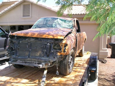 1986 Toyota Hilux - A heavily damaged, rust-colored pickup truck is loaded onto a trailer in front of a residential garage. The truck's front end is missing, a…