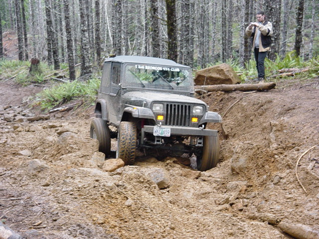 Jeep Wrangler driving on a rocky, muddy trail in a forest. A person stands on a ridge taking a photo