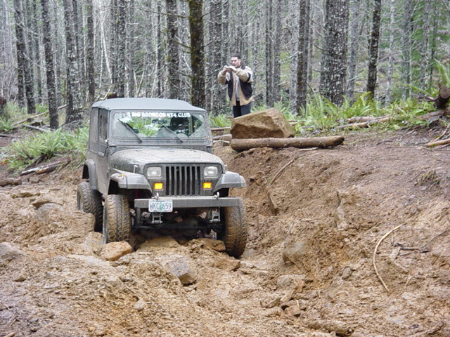 Gray Jeep Wrangler on a muddy, rocky trail with a person standing in the background