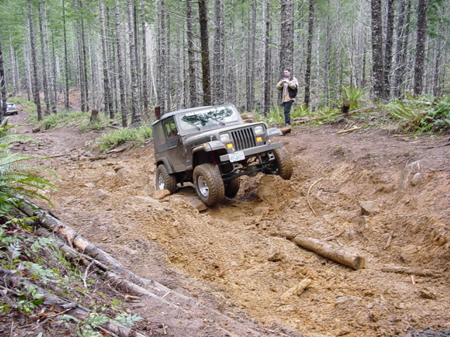 Jeep Wrangler climbing a muddy hill in a forest. A person stands on the bank watching