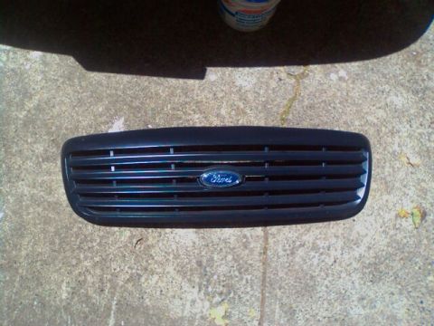 2000 Ford Crown Victoria - A black Ford grille with horizontal slats and a blue oval emblem is shown lying on a concrete surface.