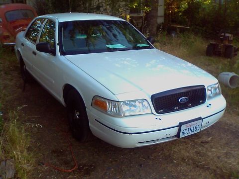 2000 Ford Crown Victoria - A white 2000 Ford Crown Victoria sedan is parked in a yard, with a red car visible behind it.