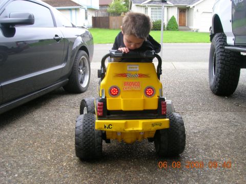 1996 Ford Bronco - A child sits in a yellow toy Bronco, parked next to a dark car and a large truck.