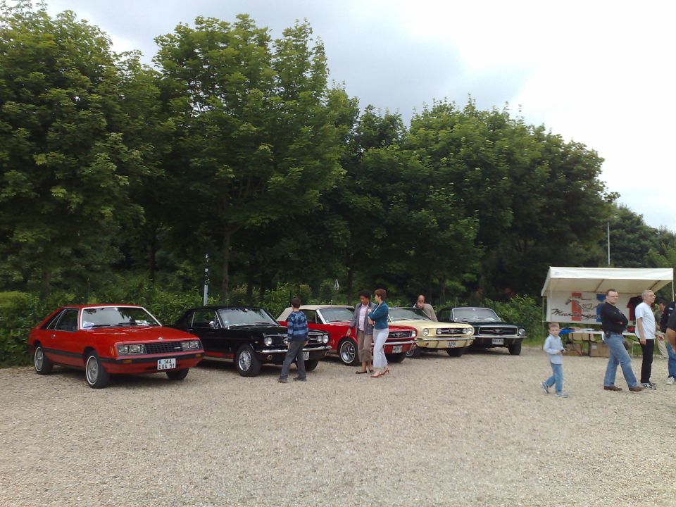 Red, black, and yellow Ford Mustangs parked in a gravel lot with trees and a tent in the background