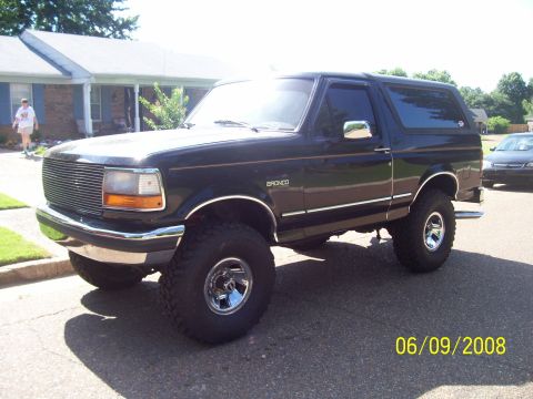 1994 Ford Bronco - A dark-colored Ford Bronco is parked on a residential street, showing its lifted stance and chrome accents.