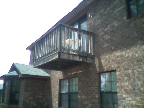 1992 Ford F-150 - Exterior view looking up at a second-story wood balcony deck cantilevered off a brown brick house, with a sliding glass door and shingled r…