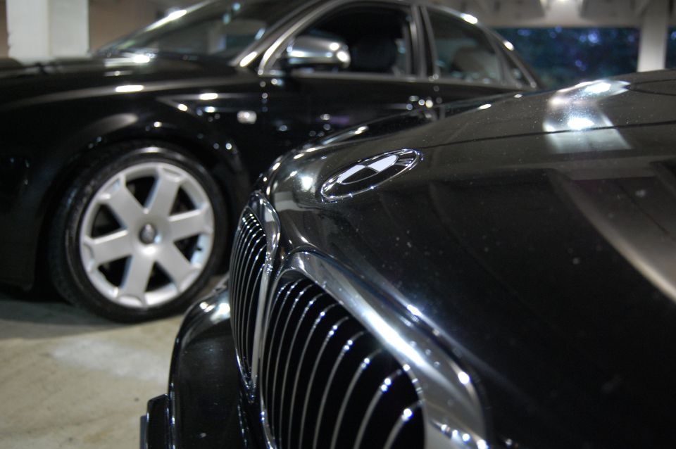 Close-up of a car grille and hood emblem, with another black car and its wheel visible behind it