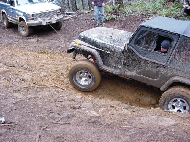 Black Jeep Wrangler stuck in a muddy ditch, with a white truck in the background pulling it