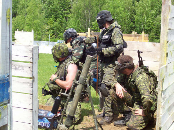 paintball players in camouflage gear, one with a mortar, in a wooded area with wooden barriers
