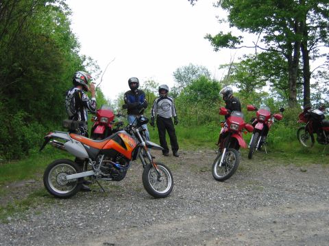 1985 Ford LTD Crown Victoria - Motorcyclists and their dirt bikes gathered on a gravel road surrounded by trees.