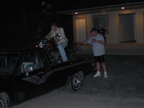 1985 Ford LTD Crown Victoria - A motorcycle is being loaded onto the bed of a black pickup truck as people stand nearby.