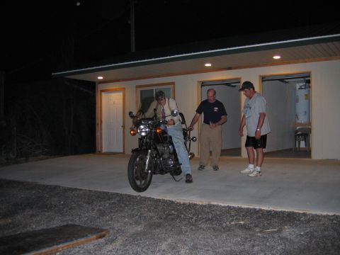1985 Ford LTD Crown Victoria - A man sits on a motorcycle in front of a garage as two other men watch.