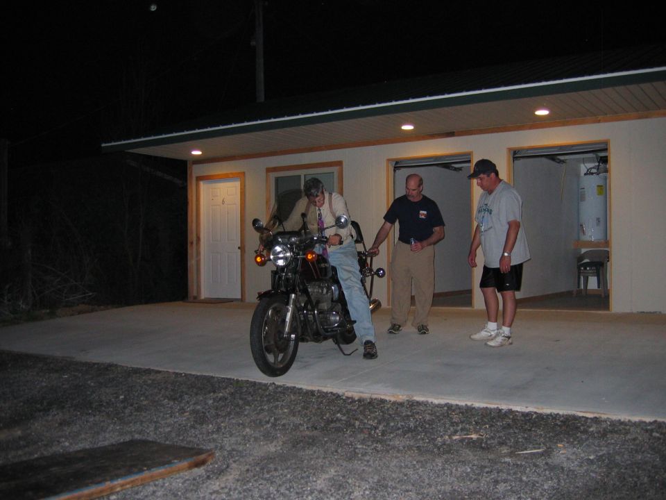 Man on a motorcycle in front of a garage with two men standing nearby