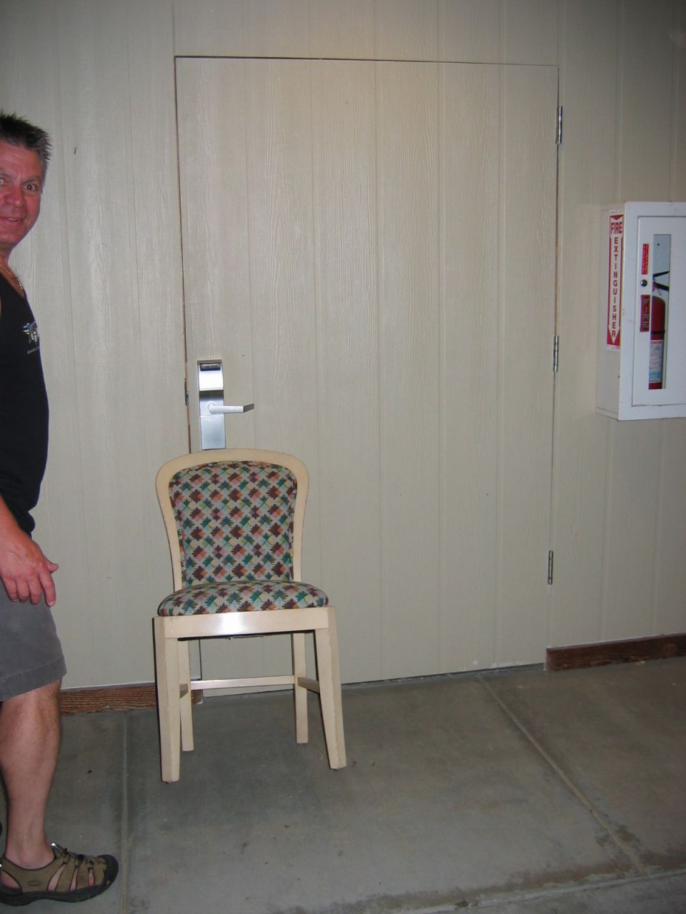 Man in black tank top and shorts stands left of a chair in front of a closed door. Fire extinguisher mounted on wall right