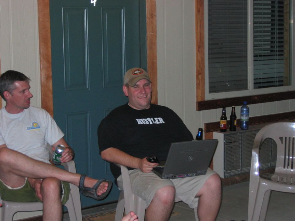 Man in black t-shirt with 'Hustler' text sits with laptop and drinks