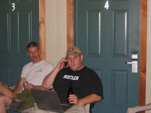1985 Ford LTD Crown Victoria - Two men sit in a hallway with doors numbered 3 and 4 behind them. One man is on his phone with a laptop and beer.