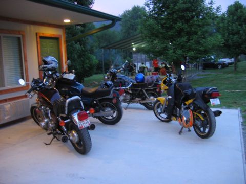 1985 Ford LTD Crown Victoria - Motorcycles are parked outside a building with people socializing in the background under a covered patio.