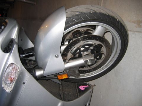 1985 Ford LTD Crown Victoria - Close-up of a silver motorcycle's rear wheel and fender assembly, showing the brake system and a decorative sticker.