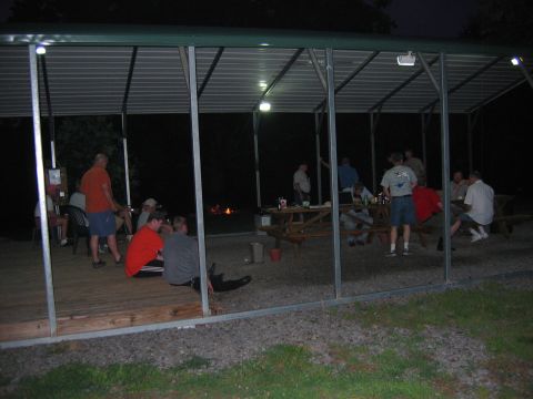 1985 Ford LTD Crown Victoria - People socialize under a covered area at night, with a campfire burning in the distance.