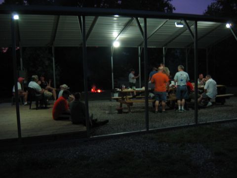 1985 Ford LTD Crown Victoria - A group of people enjoy an evening gathering around picnic tables and a bonfire under a sheltered area.