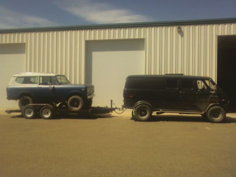 1992 Ford F-350 - A black van tows a blue SUV on a trailer in front of a large industrial building.