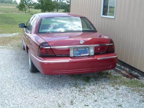 1999 Mercury Grand Marquis - The rear of a red sedan is visible, parked on a gravel surface next to a building.