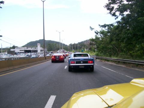 2001 Ford Mustang - A blue classic car drives down a highway, with other vehicles and a car dealership visible in the background.