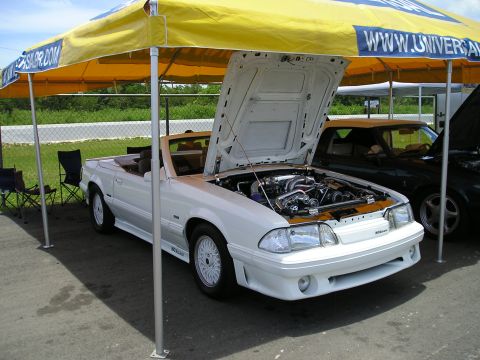 2001 Ford Mustang - A white convertible with its hood open is parked under a yellow tent next to another car.