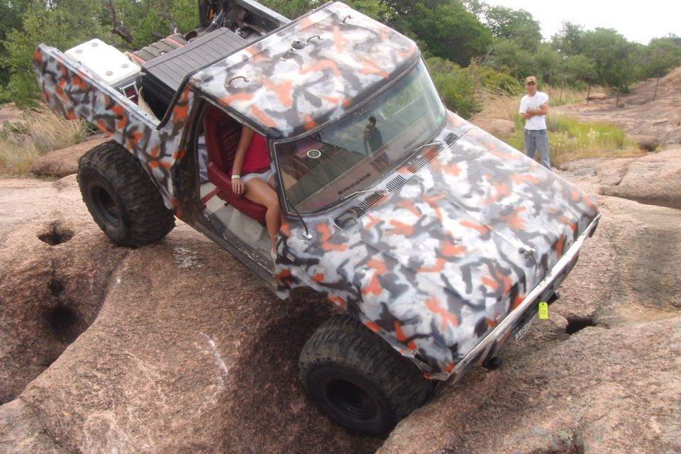 Camouflaged off-road truck climbing over large rocks with a person standing in the background