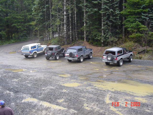 Four SUVs parked on a gravel road with trees in the background. One is a white and blue SUV, two are dark gray SUVs, and one is a silver and black SUV