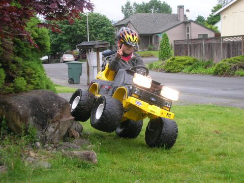 1996 Ford Bronco - A child drives a yellow toy truck over grass and rocks, giving a peace sign.