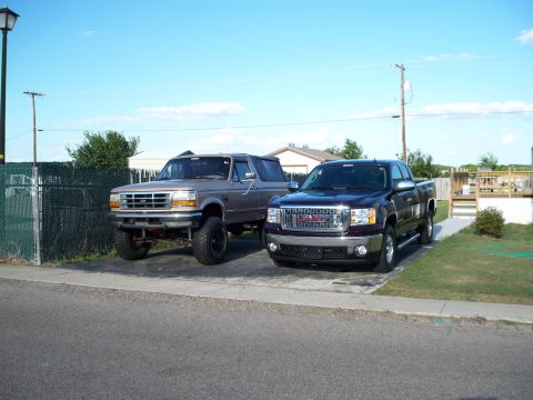 1996 Ford Bronco - Two trucks parked side by side on a residential concrete driveway under a clear sky: a lifted tan Ford two-door SUV on mud tires with its s…