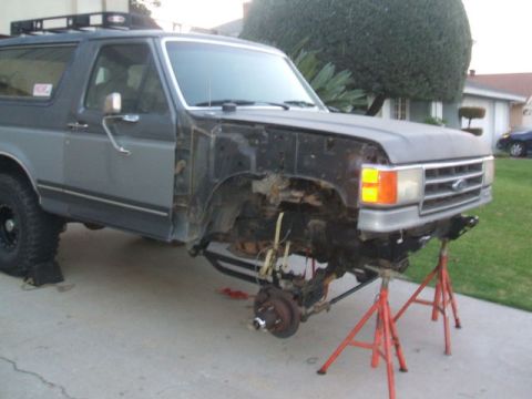 1988 Ford Bronco - Passenger-side 3/4 front view of a two-tone gray/silver full-size Ford Bronco SUV mid-teardown in a residential driveway, with the front fe…