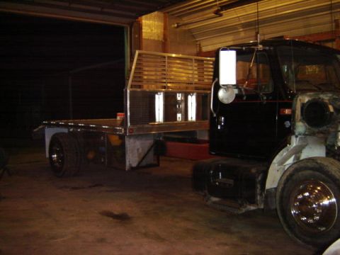 1992 Ford Bronco - A black truck chassis with a flatbed is parked indoors, showing its unfinished front end.