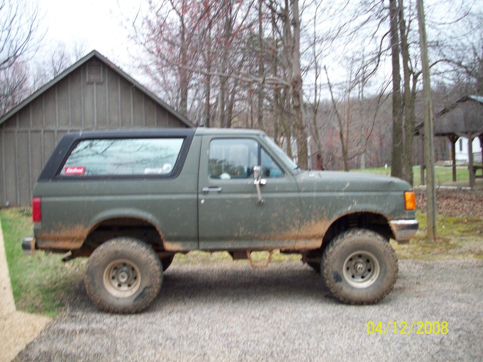 Side view of a 1987 Ford Bronco, lifted with off-road tires. It's parked in a rural setting near a wooden building