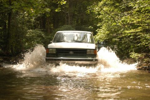 1987 Ford Bronco - A white and green 1987 Ford Bronco splashes through a shallow river surrounded by trees.