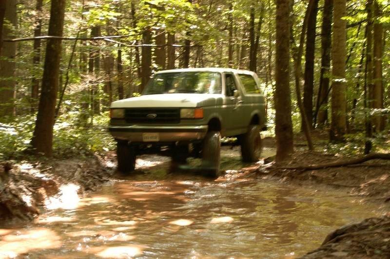 Green 1987 Ford Bronco driving through a muddy forest stream