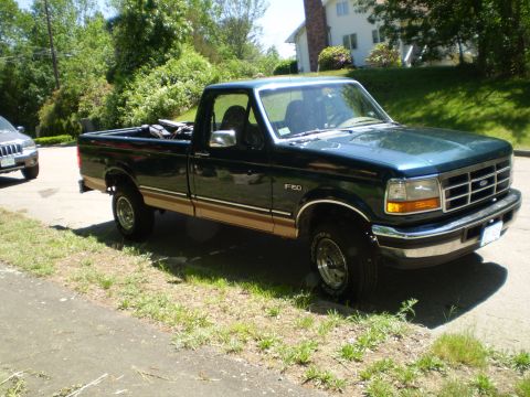 1995 Ford F-150 - A dark green 1995 Ford F-150 pickup truck is parked on the side of the road next to another vehicle.