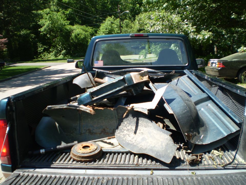 Rear view of a dark green pickup truck bed filled with various metal parts. Another car is visible in the background