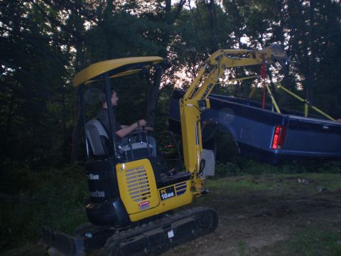 1995 Ford F-150 - A mini excavator is lifting a blue truck bed in a wooded area.
