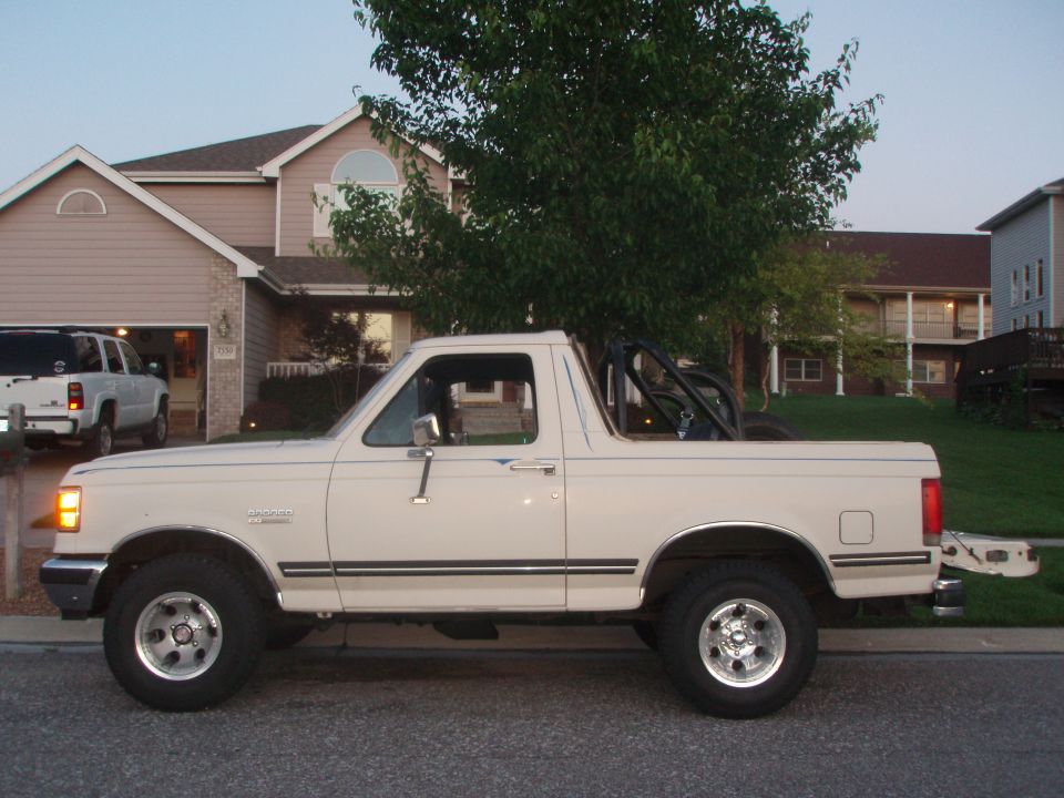 Cream-colored 1987 Ford Bronco, side view, with aftermarket roll cage and chrome wheels