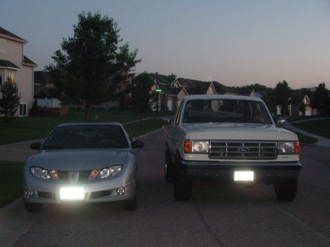 1987 Ford Bronco - Two vehicles, a Ford truck and a Pontiac, parked on a residential street at dusk.