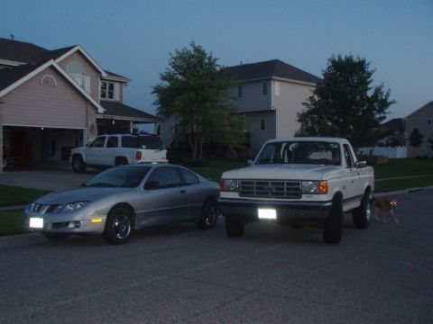 1987 Ford Bronco - Two vehicles are parked on a residential street as the sky darkens, with a dog nearby.