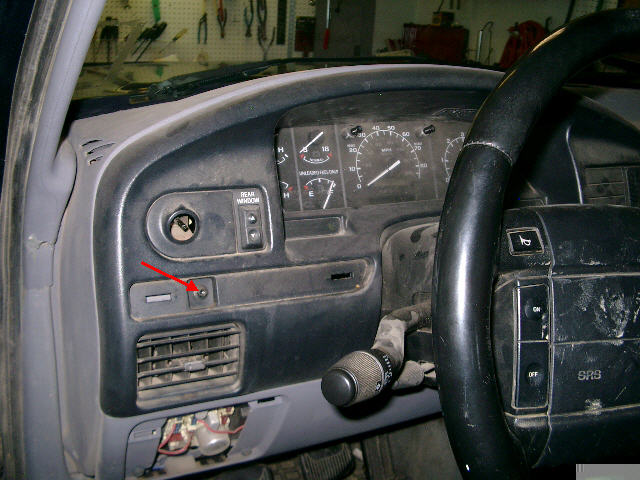 Gray Ford dash with red arrow marking bezel screw below headlight and rear window switches