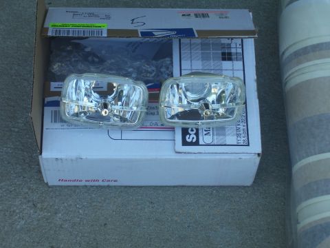 1979 Chevrolet Camaro - Two clear plastic car headlights with chrome accents are placed on a cardboard box, with a roll of striped material to the right.